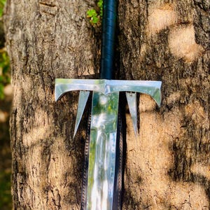 May include: A silver sword with a black leather handle and a decorative cross guard. The sword is leaning against a tree trunk.