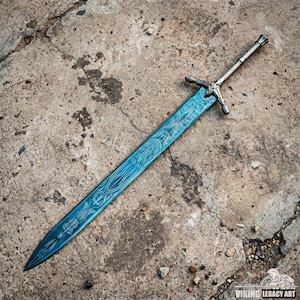 May include: A long, ornate sword with a blue blade and silver hilt. The blade features intricate etched patterns. The hilt has a crossguard design. The sword is displayed on a textured concrete surface. The image is well-lit, highlighting the details of the weapon.