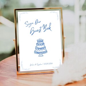 May include: A white sign with blue text and a blue illustration of a wedding cake. The sign reads "Sign Our Guest Book" and "Julia & Sophia 10.08.2016".