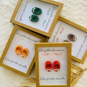 May include: Four framed baby booties in various colors: green, orange, brown, and red. Each frame has the text "Des petits chaussons..." and "Une grande nouvelle". The frames are made of light brown wood.