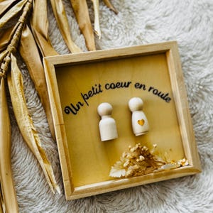 Op de afbeelding: Houten kunstwerk met een lichtbruine achtergrond. Twee houten peg dolls bevinden zich in de lijst. Eén pop heeft een hartje erop geschilderd. De tekst "Un petit coeur en route" is in zwart cursief geschreven. Gedroogde bloemen staan onderaan.