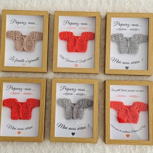 May include: A collection of framed baby announcements featuring miniature crocheted sweaters in various colors, including green, pink, red, and gray. Each frame contains text in French, with a heart symbol. The frames are displayed on a light-colored surface.