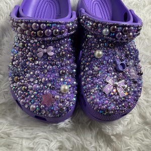 May include: A pair of purple clogs decorated with rhinestones, pearls, and bows. The clogs are covered in a sparkly, iridescent material.