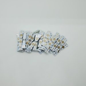 May include: A collection of small, white, rectangular printed circuit boards arranged in a pile. Each board features a central circular gold-colored contact pad with radiating lines, surrounded by smaller gold connection points and white solder mask.