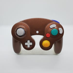 May include: A metallic brown video game controller featuring a large green A button, a red B button, a yellow C-stick, a white D-pad, and a grey analogue stick. The controller includes a blue Z button on the top right and white X and Y buttons.