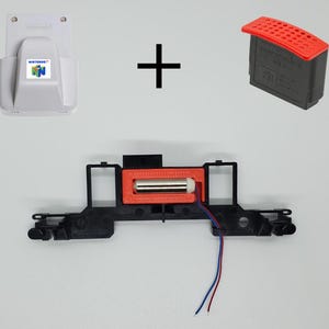 May include: A white Nintendo 64 expansion pak cover, a black and red memory expansion pak, and a black plastic internal component featuring a red 3D-printed housing with a silver cylindrical motor and two thin red and blue electrical wires.