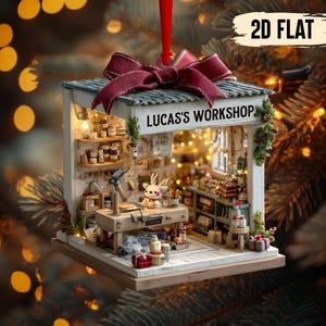 Puede incluir: Adorno navideño en miniatura con forma de taller, con el texto "LUCAS'S WORKSHOP" sobre la entrada. El adorno presenta un lazo rojo, un banco de trabajo y estanterías llenas de objetos en miniatura. El fondo está borroso con luces bokeh.