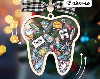 May include: A tooth-shaped shaker ornament with a black and white buffalo plaid ribbon. The interior features dental and medical illustrations, including a name tag reading Emily Nurse, a tooth labeled 2024, and various tools. It contains glitter.