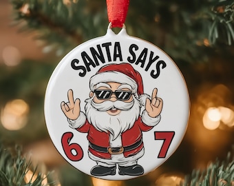 May include: A white Christmas ornament with a cartoon Santa Claus wearing sunglasses and a red suit. The ornament has the text "SANTA SAYS" and the numbers "6" and "7". A red ribbon is attached for hanging.