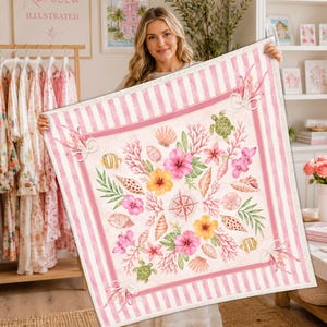 May include: A square, pink and white striped blanket with a central design featuring seashells, coral, flowers, and a compass rose. The blanket has a border of pink stripes and is held up by a person.