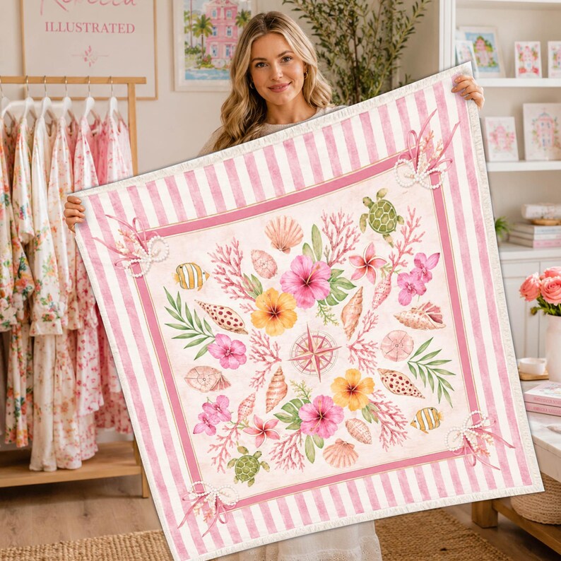 May include: A square, pink and white striped blanket with a central design featuring seashells, coral, flowers, and a compass rose. The blanket has a border of pink stripes and is held up by a person.