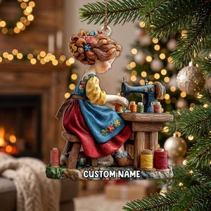 May include: A hanging ornament featuring a figure with auburn hair styled with colourful flowers, wearing a blue apron over a yellow shirt, sitting on a wooden stool at a sewing machine. The base includes spools of thread and the text CUSTOM NAME.