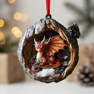 May include: A round hanging ornament featuring a red dragon with spread wings nestled inside a textured, hollowed-out tree trunk. The base of the ornament displays the name William in script, surrounded by small red berries and white snow accents.