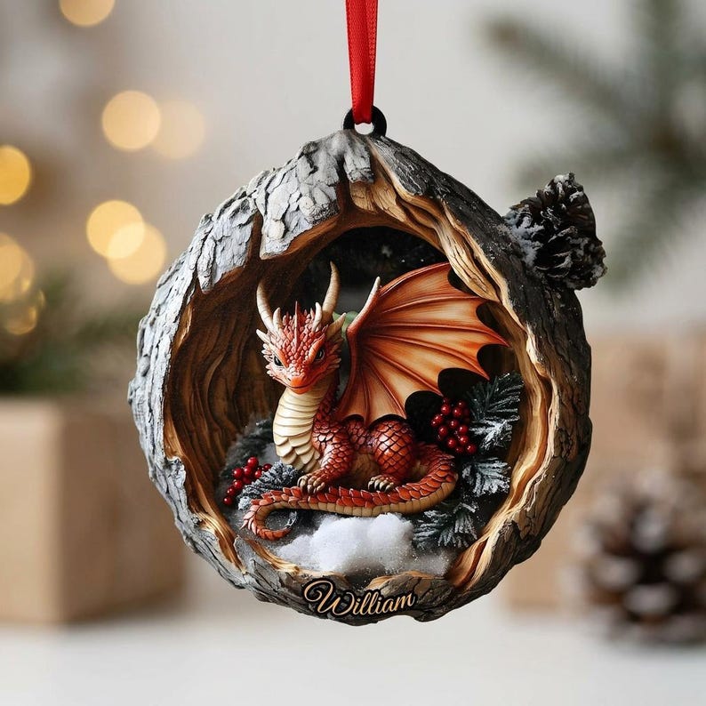 May include: A round hanging ornament featuring a red dragon with spread wings nestled inside a textured, hollowed-out tree trunk. The base of the ornament displays the name William in script, surrounded by small red berries and white snow accents.