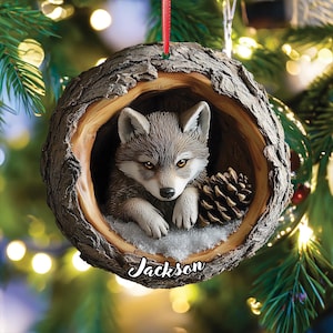 May include: A round, tree-trunk-shaped Christmas ornament featuring a sculpted wolf pup resting inside a hollow log with a pinecone and a dusting of snow. The name Jackson is printed in white script at the bottom of the ornament, which hangs by a red ribbon.