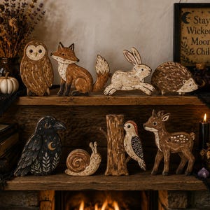 May include: A collection of hand-painted wooden animal figurines including an owl, fox, rabbit, hedgehog, crow with moon and leaf motifs, snail, woodpecker on a tree trunk, and a fawn, arranged on a wooden mantelpiece with decorative items.