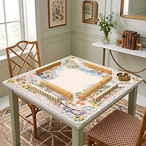 May include: A square mahjong table with a floral and landscape design. The table is set with wooden mahjong tiles. The table is surrounded by two bamboo chairs and a patterned rug. The table is approximately 36 inches square.