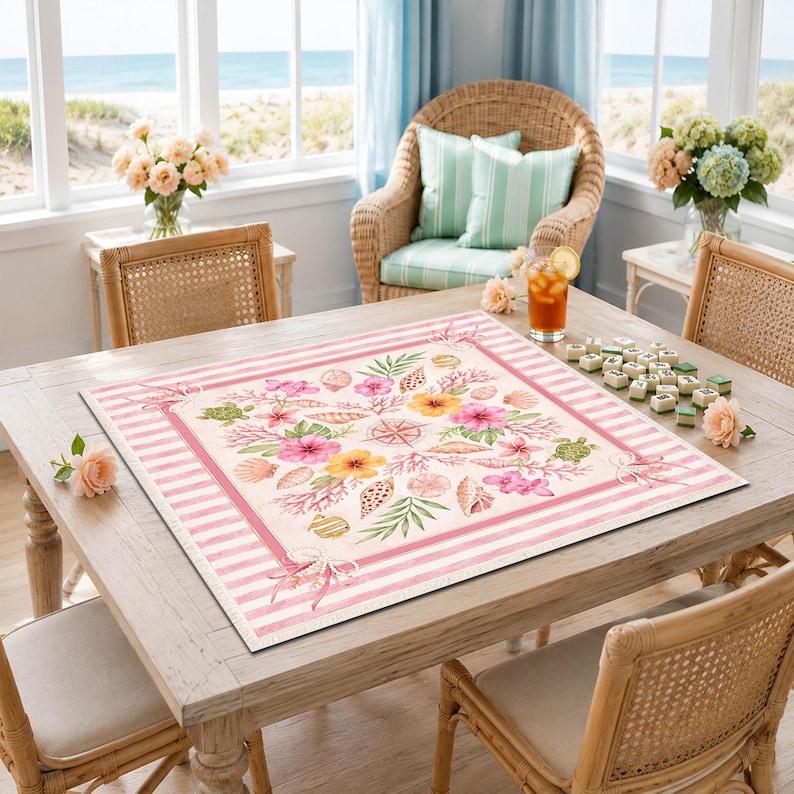 May include: A square, pink and white striped placemat with a floral and seashell design. The placemat features pink hibiscus flowers, seashells, and green leaves. The placemat is on a light wood table.