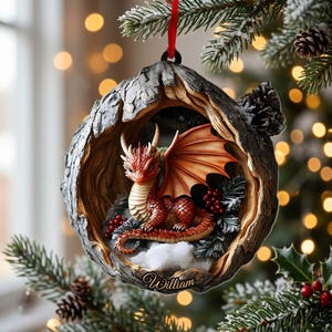 May include: A round Christmas tree ornament featuring a sculpted red dragon resting inside a hollowed-out tree trunk. The dragon has orange wings and scales. The base of the ornament is inscribed with the name William in a cursive script.