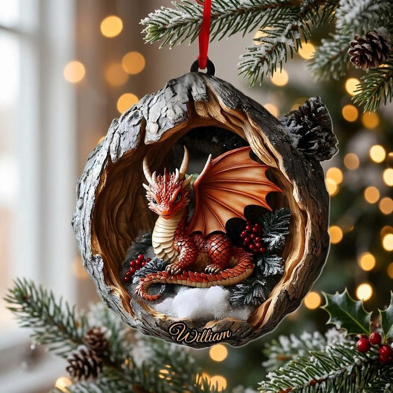 May include: A round Christmas tree ornament featuring a sculpted red dragon resting inside a hollowed-out tree trunk. The dragon has orange wings and scales. The base of the ornament is inscribed with the name William in a cursive script.