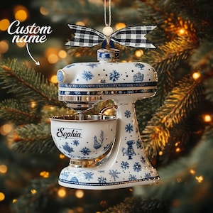 May include: A white and blue ceramic stand mixer Christmas ornament. The ornament features a blue and white snowflake design, gold accents, and the name "Sophia" on the bowl. It is topped with a black and white checkered bow.