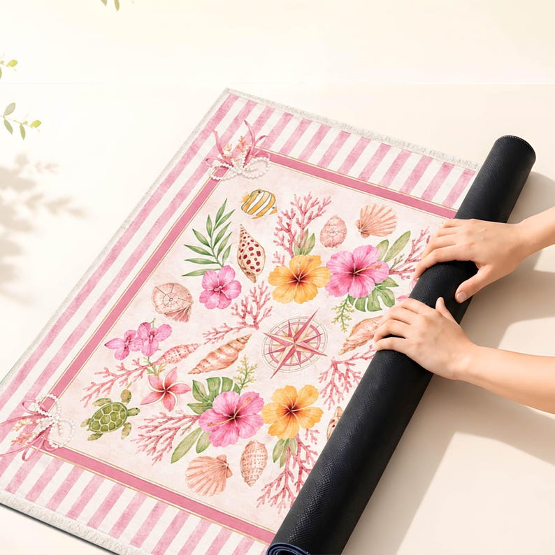 May include: A rolled-up yoga mat with a coastal design. The mat features a cream background with pink stripes and a central design of seashells, flowers, and a compass rose. The mat is being rolled up by a person.
