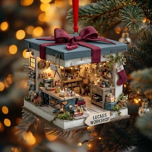 May include: A miniature workshop ornament shaped like a gift box with a maroon ribbon on top. The interior features a workbench, shelves with tools, and a sign that reads LUCAS'S WORKSHOP. A maroon stocking hangs on the side of the white structure.