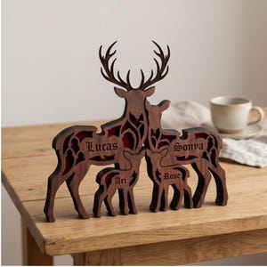 Puede incluir: Escultura de madera de una familia de ciervos con los nombres "Lucas", "Sonya", "Ari" y "Rose" grabados. Los ciervos marrones tienen un interior rojo y están sobre una mesa de madera. Objeto decorativo.