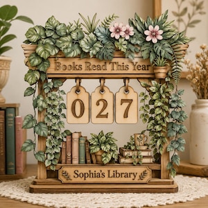 May include: Wooden bookshelf-style calendar with the text "Books Read This Year" and "Sophia's Library." The calendar displays the number "027" on hanging wooden tags. The design includes faux greenery, flowers, and miniature books.