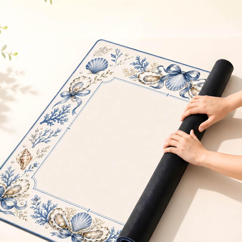 Puede incluir: Una alfombrilla rectangular con un centro de color crema y un borde decorativo con ilustraciones en azul y beige de conchas marinas, corales y cintas. La alfombrilla tiene una base negra s&oacute;lida y est&aacute; parcialmente enrollada para mostrar su textura.