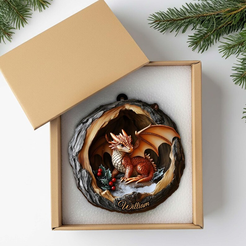 May include: A round, tree-trunk-shaped ornament featuring a small, orange-scaled dragon inside a hollow, with red holly berries and the name William inscribed at the bottom, presented in a square cardboard gift box with a matching lid.