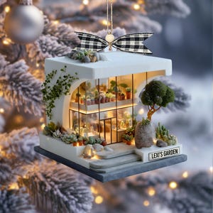 Puede incluir: Un adorno de invernadero en miniatura de dos pisos en color blanco con paredes de cristal, estantes interiores con suculentas en macetas, un árbol pequeño y detalles de piedra. Un lazo de cuadros blanco y negro está sujeto en la parte superior. Un letrero dice LEXI'S GARDEN.