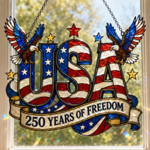 Könnte beinhalten: Ein Buntglas-Fensterschmuck mit den Buchstaben "USA" in Rot, Weiß und Blau, mit Sternen und Adlern. Auf dem Banner steht "250 YEARS OF FREEDOM". Das Stück ist zum Aufhängen mit einer Kette konzipiert.