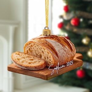 May include: A hanging ornament shaped like a loaf of sourdough bread with one slice resting on a wooden cutting board. The ornament features a gold cap and string, with the name Sophia and the year 2025 written in white cursive script on the board.