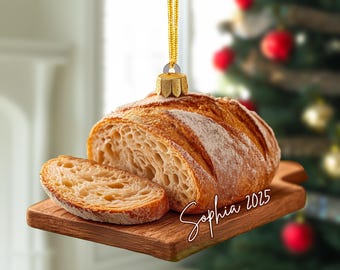 May include: A hanging ornament shaped like a loaf of sourdough bread with one slice resting on a wooden cutting board. The ornament features a gold cap and string, with the name Sophia and the year 2025 written in white cursive script on the board.