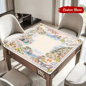May include: A square table cover featuring a watercolor design of a coastal town with buildings, bridges, and flowers. The cover has a cream-colored center and a decorative border. The text "Custom Name" is at the top.