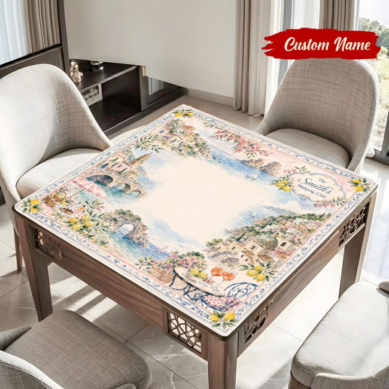 May include: A square table cover featuring a watercolor design of a coastal town with buildings, bridges, and flowers. The cover has a cream-colored center and a decorative border. The text "Custom Name" is at the top.