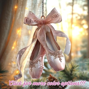 May include: A pair of pale pink ballet slippers hanging from a chain, adorned with a matching ribbon featuring the name Olivia and the year 2025. The slippers are decorated with intricate gold floral embellishments on the front toe sections.