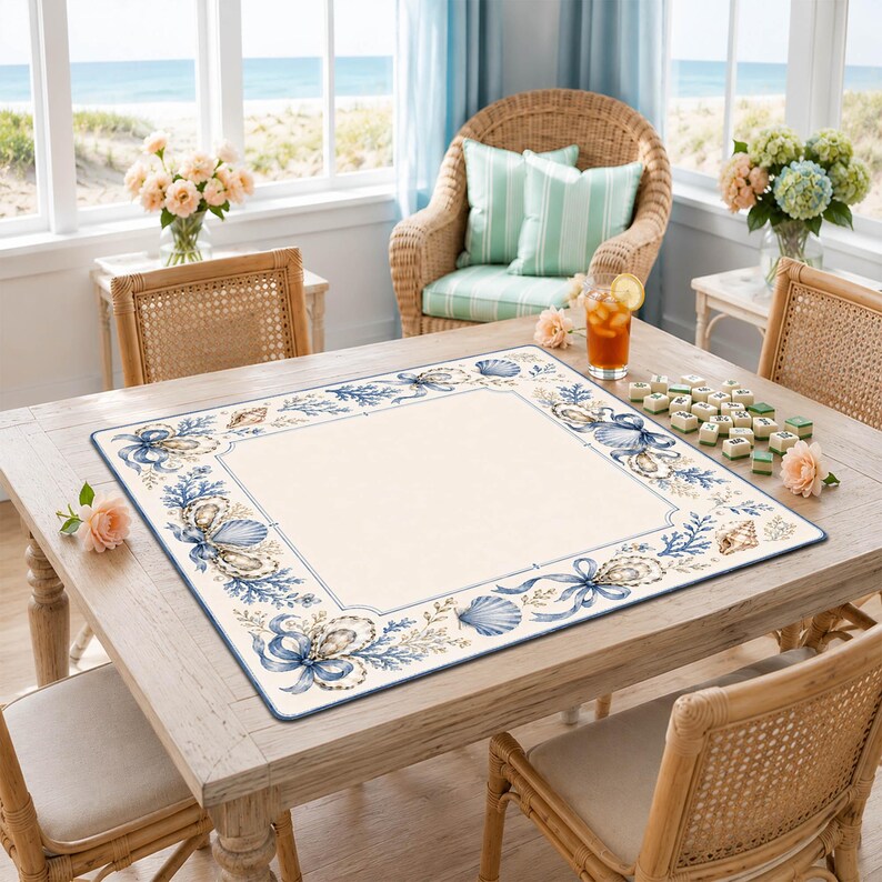 Puede incluir: Un mantel individual cuadrado con un centro color crema y un borde decorativo con ilustraciones en azul y beige de conchas marinas, corales y cintas. El mantel est&aacute; sobre una mesa de madera con fichas de juego y un vaso de t&eacute; helado.