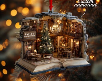 May include: A three-dimensional hanging ornament shaped like an open book, featuring a detailed miniature bookstore interior with a Christmas tree, a chandelier, bookshelves, and a sign that reads EMILY'S BOOKSTORE, suspended by a red ribbon.