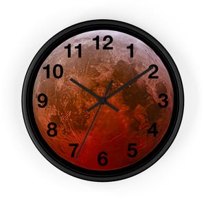 Puede incluir: Un reloj de pared redondo con marco negro y una imagen roja y naranja de Marte en el centro. La esfera del reloj tiene números negros del 1 al 12.