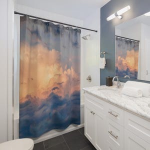 May include: A bathroom with a shower curtain featuring a sunset sky with fluffy clouds. The shower curtain is a light blue color with orange and pink clouds. The bathroom has a white vanity with a double sink and a large mirror.