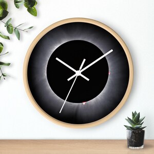 Puede incluir: Un reloj de pared redondo con una imagen en blanco y negro de un eclipse solar. El reloj tiene manecillas blancas y un marco de madera.