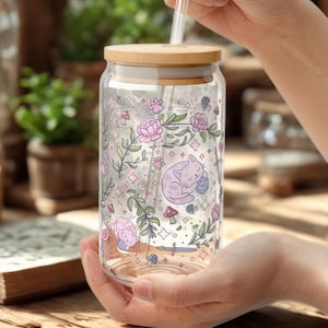 Könnte beinhalten: Ein klares Glas mit einem Holzdeckel und einem durchsichtigen Strohhalm. Der Becher ist mit einem Blumen- und Katzenmuster in Rosa und Lila verziert. Der Becher wird von einer Person gehalten.