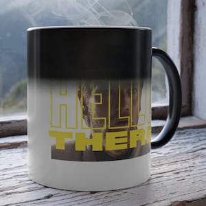 Op de afbeelding: Een zwart-witte warmtegevoelige mok met de tekst "HELLO THERE" in geel. De mok toont een portret van een man. Stoom stijgt op uit de mok, wat duidt op een warme drank. De mok staat op een verweerd houten oppervlak.