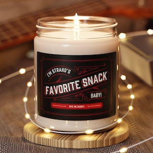 May include: A lit candle in a glass jar with a black and red label that reads "I'M STRAHO'S FAVORITE SNACK BABY! BITE ME DADDY!". The candle sits on a round wooden coaster, with a guitar and book in the background.
