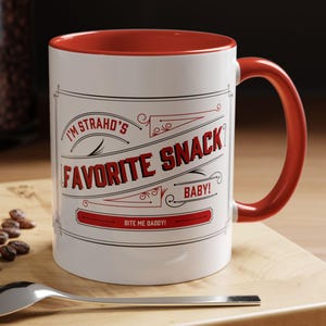 Puede incluir: Taza de cerámica blanca con interior y asa rojos. La taza presenta el texto "I'M STRAHD'S FAVORITE SNACK BABY!" en rojo y negro, con "BITE ME DADDY!" debajo. Se ven una cuchara y un macaron.