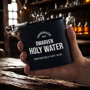 Op de afbeelding: Een zwarte heupfles met een scharnierende dop en een zilverkleurige bovenkant. De fles heeft de witte tekst "HANDMADE 1001 DWARVEN HOLY WATER (DEFINITELY NOT ALE)". De fles wordt in een hand gehouden.