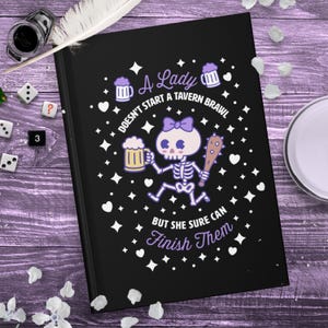 Op de afbeelding: Zwart notitieboek met de tekst "A Lady Doesn't Start a Tavern Brawl But She Sure Can Finish Them." De cover toont een skelet met een strik, bier en knots, met witte sterren en harten.