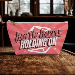 May include: A pink zippered pouch with the text "BAG OF BARELY HOLDING ON" in a retro font. The design features a pink radial pattern and a white geometric shape. The zipper is gold-colored.
